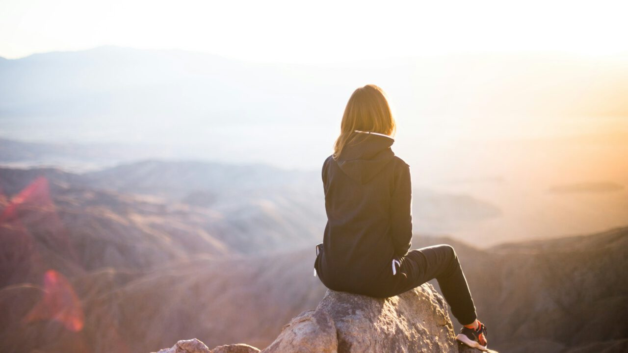 Frau sitzt auf einem Felsen und blickt in eine weite Berglandschaft bei Sonnenaufgang – Symbol für Klarheit, Selbstreflexion und neue Perspektiven im astrologischen Life Coaching und beruflichen Coaching.