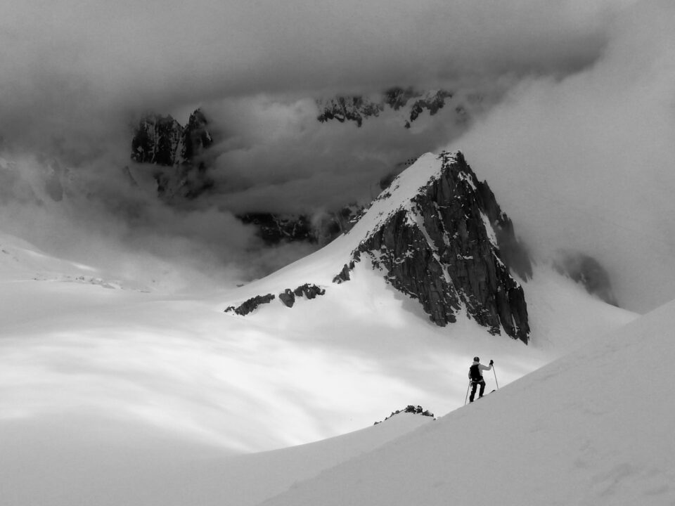 Person erklimmt mit Skistöcken eine schneebedeckte Berglandschaft, umgeben von dramatischen Wolken und schroffen Gipfeln – symbolisch für Herausforderungen, Zielsetzung und persönliche Entwicklung durch astrologisches Coaching.