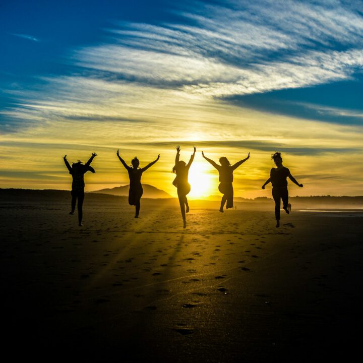 Gruppe von Menschen springt freudig am Strand bei Sonnenuntergang – Symbol für Gemeinschaft, Lebensfreude und positive Transformation durch astrologisches Coaching.