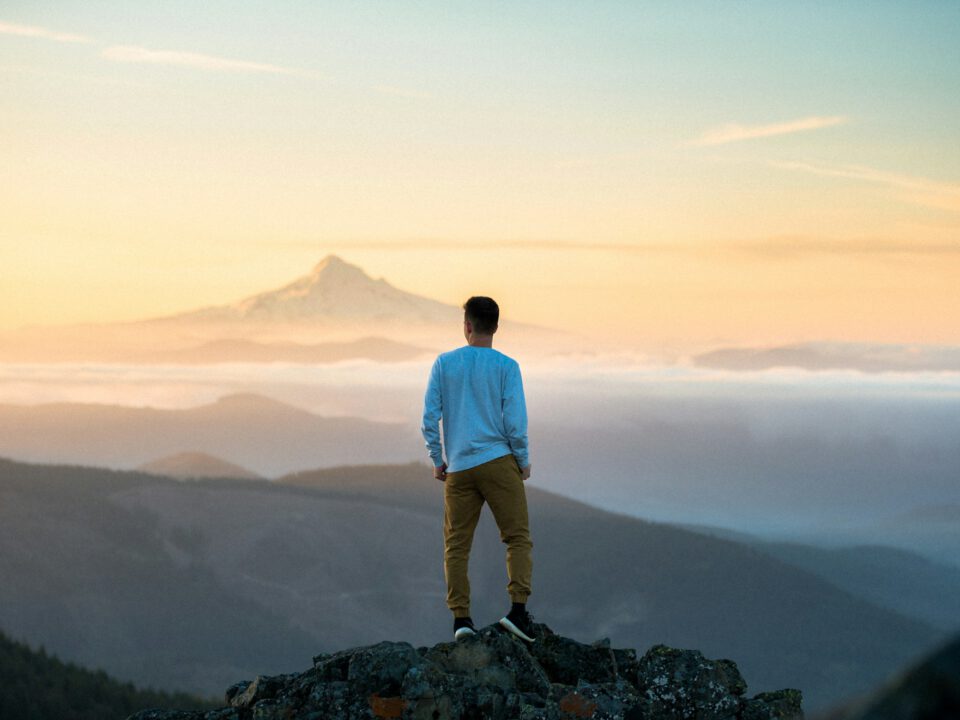 Mann steht auf einem Felsen und blickt in eine weite Berglandschaft mit Blick auf einen schneebedeckten Gipfel bei Sonnenaufgang – Symbol für Klarheit, Zielsetzung und Perspektiven im astrologischen Life Coaching und beruflicher Weiterentwicklung.